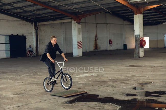 Vista lateral de um jovem caucasiano vestido de preto montando na roda traseira de uma bicicleta BMX enquanto pratica truques em um armazém abandonado — Fotografia de Stock