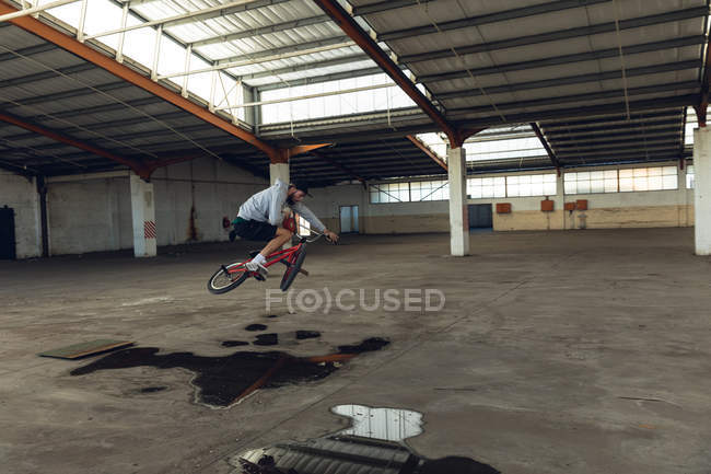 Vue latérale d'un jeune homme caucasien en vélo BMX, sautant du sol et tournant le guidon, tout en pratiquant des tours dans un entrepôt abandonné — Photo de stock