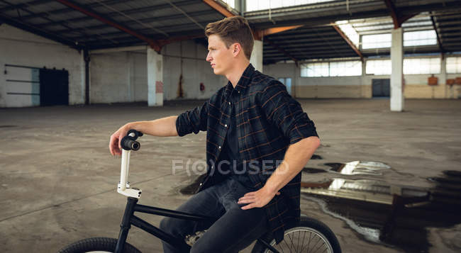 Vista lateral de perto de um jovem caucasiano vestido de preto, sentado em uma bicicleta BMX e olhando para longe em um armazém abandonado — Fotografia de Stock