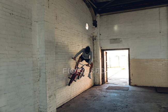 Vista frontal de um jovem caucasiano que anda de bicicleta BMX em um corredor vazio em um armazém abandonado — Fotografia de Stock