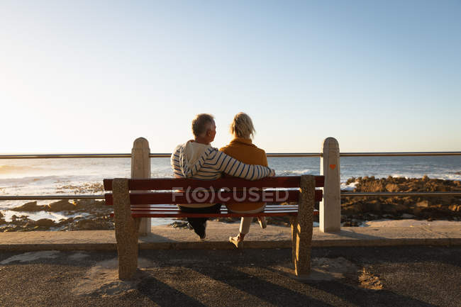 Vista trasera de cerca de un hombre y una mujer caucásicos maduros sentados en un banco y admirando la vista por el mar al atardecer - foto de stock