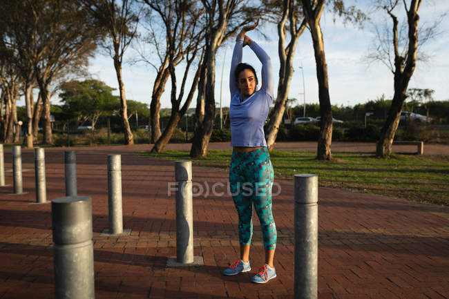 Front view of a young Caucasian woman wearing sports clothes raising her arms and stretching while working out in a park — Stock Photo