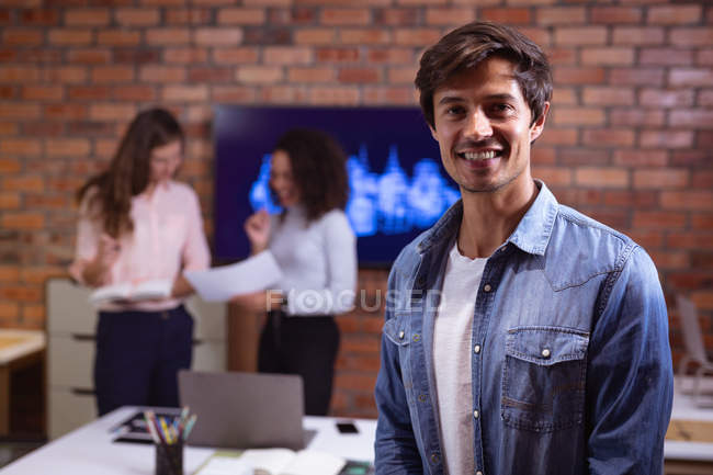 Porträt eines jungen kaukasischen Mannes, der im Büro eines kreativen Unternehmens arbeitet und in die Kamera lächelt, während zwei Kolleginnen im Hintergrund miteinander reden — Stockfoto