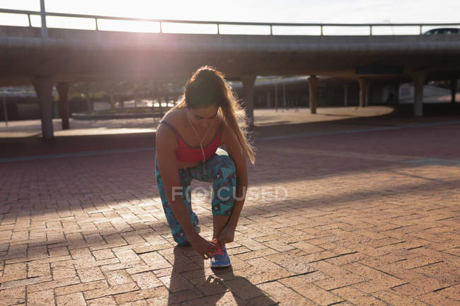 Front view of a young Caucasian woman wearing sports clothes kneeling and tying her shoe while listening to music on earphones before working out on a sunny day in a park — Stock Photo