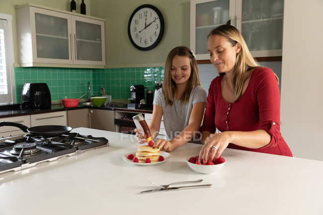 Вид спереди на молодую кавказскую женщину, которая печет блинчики дома на кухне со своей дочерью-подростком — стоковое фото