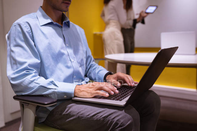 Vista lateral seção média de um jovem caucasiano sentado usando um computador portátil de joelhos em uma sala de reuniões em um negócio criativo, com dois colegas de pé e falando em segundo plano — Fotografia de Stock