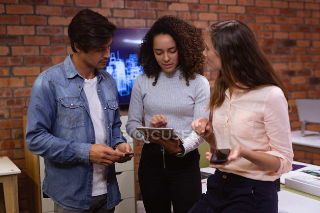 Вид спереди молодая женщина смешанной расы и молодая кавказская женщина и мужчина, стоящие и смотрящие на планшетный компьютер вместе и разговаривающие в офисе творческого бизнеса — стоковое фото