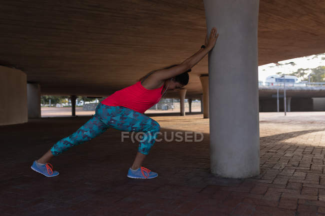 Vue latérale d'une jeune femme caucasienne portant des vêtements de sport poussant contre un poteau sous un pont et s'étirant pendant une séance d'entraînement dans un parc — Photo de stock