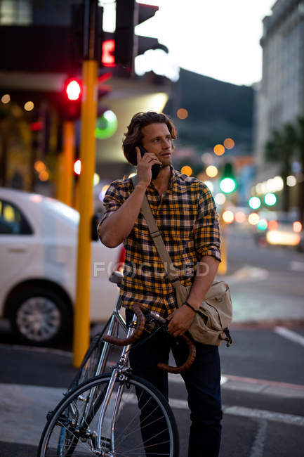 Front view close up of a young Caucasian man holding a bike and talking on a phone standing in a busy city street in the evening — Stock Photo