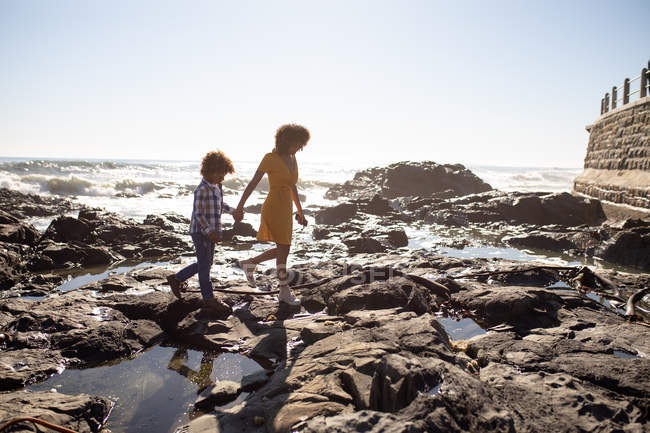 Vista lateral de uma jovem mulher de raça mista e seu filho pré-adolescente desfrutando de tempo juntos junto ao mar, de mãos dadas e andando em um dia ensolarado — Fotografia de Stock