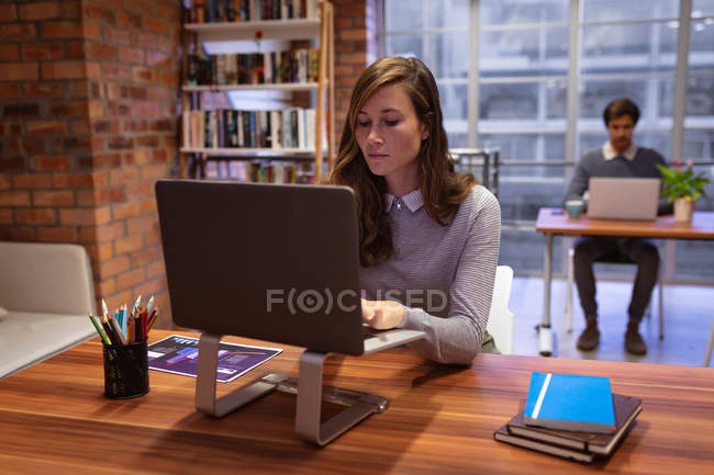 Front view close up of a young Caucasian woman working in the office of a creative business sitting at a desk using a laptop computer, with a male colleague working at a desk in the background — Stock Photo