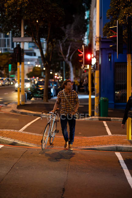Vista frontal de un joven caucásico caminando por la calle con una bicicleta por la noche - foto de stock
