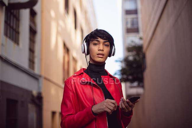 Retrato de un joven transgénero mestizo de moda en la calle, usando un smartphone con auriculares - foto de stock