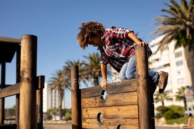 Vista laterale di un ragazzo pre-adolescente di razza mista che gioca in un parco giochi, arrampicandosi su una cornice di legno in una giornata di sole con palme ed edifici sullo sfondo — Foto stock