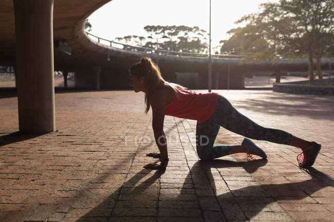 Vista lateral de uma jovem caucasiana vestindo roupas esportivas ajoelhadas e alongadas durante um treino em um parque, iluminadas pela luz solar — Fotografia de Stock