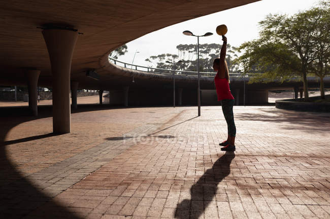 Side view of a young Caucasian woman wearing sports clothes swinging a kettlebell weight over her head during a workout on a sunny day in a park — Stock Photo