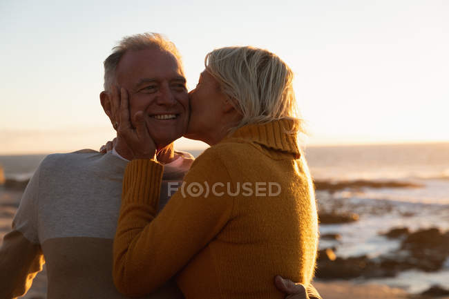 Вид сбоку: взрослый кавказский мужчина и женщина обнимаются и целуются у моря на закате — стоковое фото