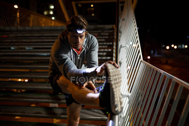 Vue de face gros plan d'un jeune homme caucasien s'étirant sur des marches dans la rue pendant son entraînement en fin de soirée avec une lampe frontale allumée — Photo de stock