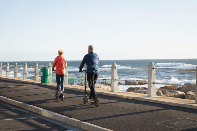 Vista trasera de un hombre y una mujer caucásicos maduros montando scooters junto al mar - foto de stock