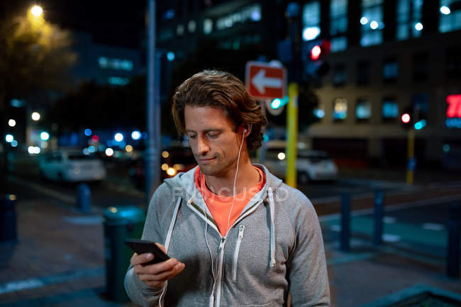 Vista frontal de cerca de un joven caucásico usando un teléfono inteligente con auriculares en la calle durante su entrenamiento nocturno - foto de stock