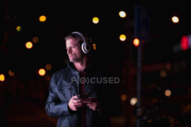 Vue de face d'un jeune homme caucasien debout dans une rue la nuit écoutant de la musique avec un casque allumé, tenant un smartphone — Photo de stock