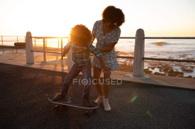 Вид спереду змішаної раси жінка і її до-підліток син насолоджується часом разом біля моря, мама допомагає синові кататися на скейтборді на набережній, підсвічуючи, встановлюючи сонце — стокове фото