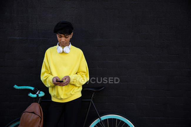 Vista frontal de un joven transexual de raza mixta de moda en la calle, mensajes de texto en el teléfono inteligente de pie junto a una bicicleta - foto de stock