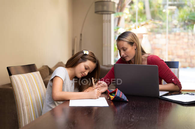 Frontansicht einer jungen kaukasischen Frau, die einen Laptop benutzt und ihrer Tochter bei den Hausaufgaben im Esszimmer hilft — Stockfoto
