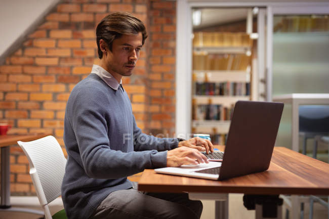 Seitenansicht eines jungen kaukasischen Mannes, der an einem Schreibtisch sitzt und einen Laptop im Büro eines kreativen Unternehmens benutzt — Stockfoto