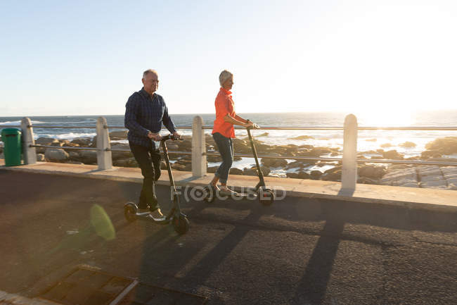 Vista lateral de um homem e uma mulher caucasianos maduros montando e scooters pelo mar no por do sol — Fotografia de Stock