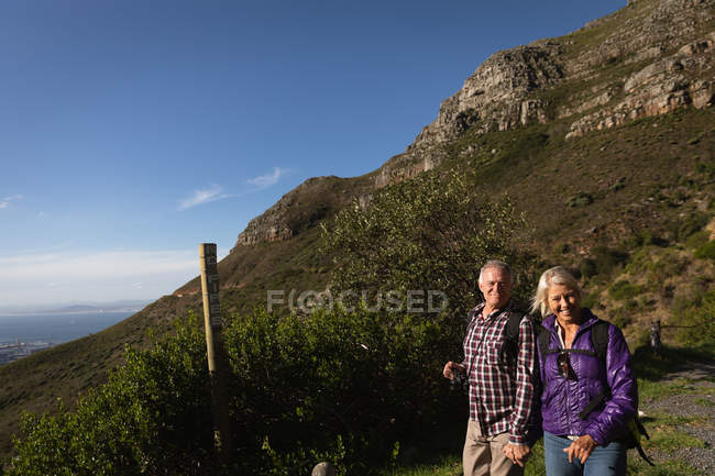 Portrait of a mature Caucasian man and woman holding hands, smiling to camera and walking in a rural setting, with mountains and a blue sky behind them — Stock Photo