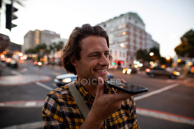 Vista frontal de cerca de un joven caucásico hablando por teléfono durante su viaje nocturno de pie en una concurrida calle de la ciudad por la noche - foto de stock