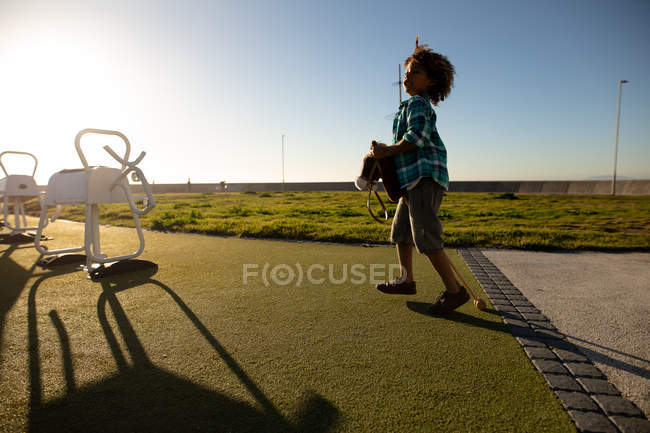 Vista lateral de un niño preadolescente de raza mixta en un parque infantil junto al mar, jugando con un caballo hobby en un día soleado - foto de stock