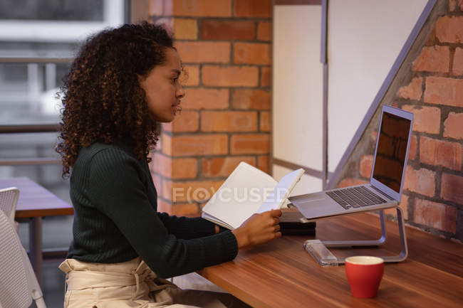 Seitenansicht einer jungen Frau mit gemischter Rasse, die im Büro eines kreativen Unternehmens arbeitet, an einem Schreibtisch sitzt, ein Notizbuch hält und auf einen Laptop blickt — Stockfoto