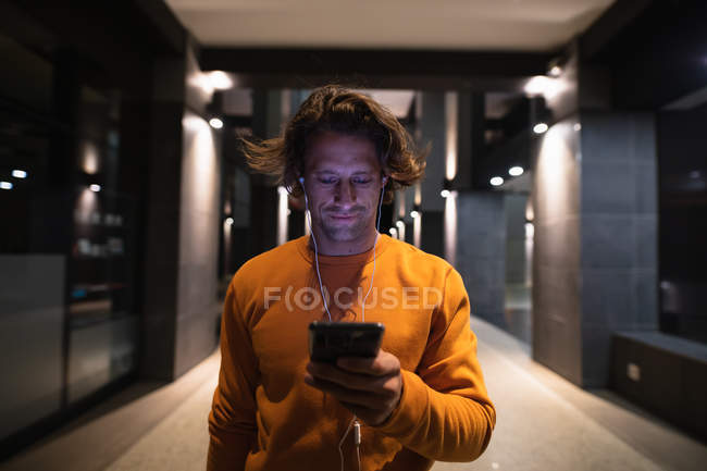Vista frontal de cerca de un joven caucásico en la calle por la noche mirando un teléfono inteligente y usando auriculares - foto de stock