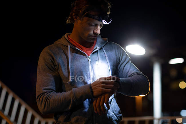 Vista frontal de cerca de un joven caucásico revisando un reloj inteligente en la calle durante su entrenamiento nocturno con un faro encendido - foto de stock