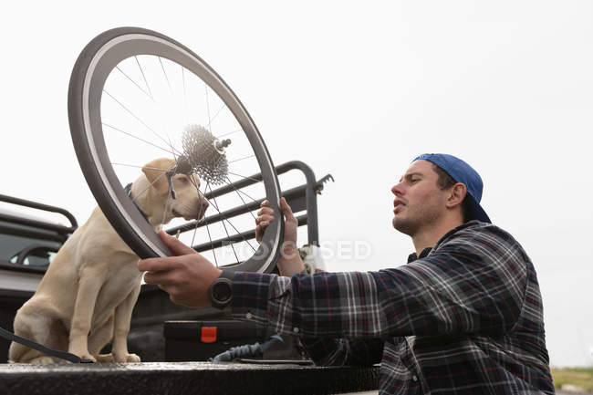 Side view close up of a young Caucasian man in a wheelchair taking a recumbent bicycle out of the back of his car to assemble it, holding a wheel with his dog watching — Stock Photo