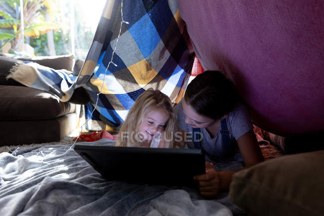Vue de face d'une jeune fille caucasienne et sa soeur entre deux en utilisant une tablette ensemble dans une tente faite de couvertures — Photo de stock