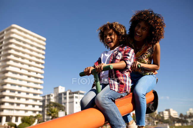 Вид спереди на молодую женщину смешанной расы и ее сына-подростка, наслаждающуюся временем вместе, играющую на детской площадке, сидящую на качелях в солнечный день со зданиями на заднем плане — стоковое фото