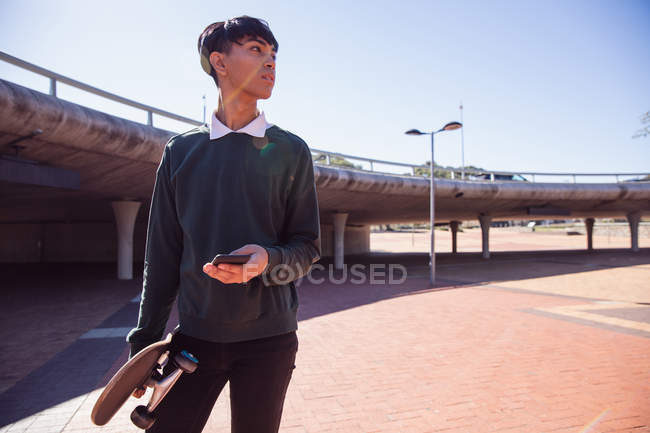 Vista frontal de um jovem elegante mestiço transexual adulto na rua, segurando um smartphone e um skate — Fotografia de Stock