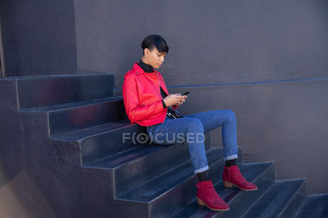 Vista lateral de un joven transexual de raza mixta de moda en la calle, sentado en escalones usando un smartphone contra una pared gris - foto de stock