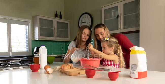 Вид спереди на молодую кавказскую женщину, готовящую вместе со своими младшими дочерьми на кухне — стоковое фото