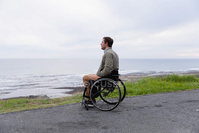 Vista lateral de un joven caucásico en silla de ruedas disfrutando de un día junto al mar, disfrutando de la vista - foto de stock