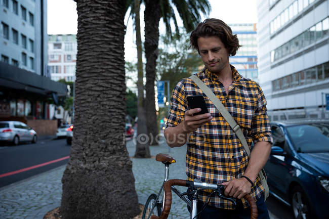 Vista frontal de um jovem caucasiano segurando uma bicicleta e usando um smartphone em uma movimentada rua urbana à noite — Fotografia de Stock