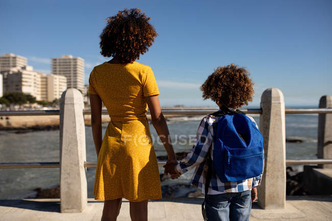 Вид спереду молода змішаної раси жінка і її попередній син насолоджуються часом разом біля моря, тримаючи руки і захоплюючись видом на сонячний день — стокове фото