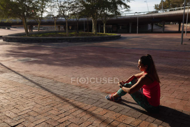 Вид сбоку на молодую кавказскую женщину в спортивной одежде, сидящую на земле и проверяющую свои умные часы во время тренировки в солнечный день в парке — стоковое фото