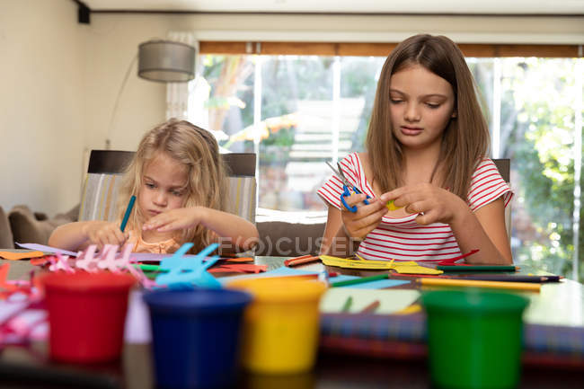 Вид спереди на юную кавказскую девушку и её младшую сестру, занимающуюся ремеслами в своей гостиной — стоковое фото