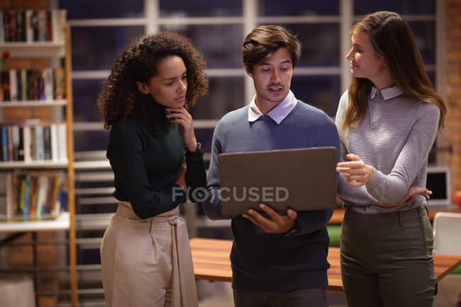 Front view close up of a young mixed race woman and a young Caucasian woman and man standing and looking at a laptop computer which the man is holding and having a discussion in the office of a creative business — Stock Photo