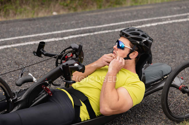 Vista lateral de cerca de un joven hombre caucásico en ropa deportiva en una bicicleta reclinada en bicicleta en un camino rural, sujetando un casco - foto de stock
