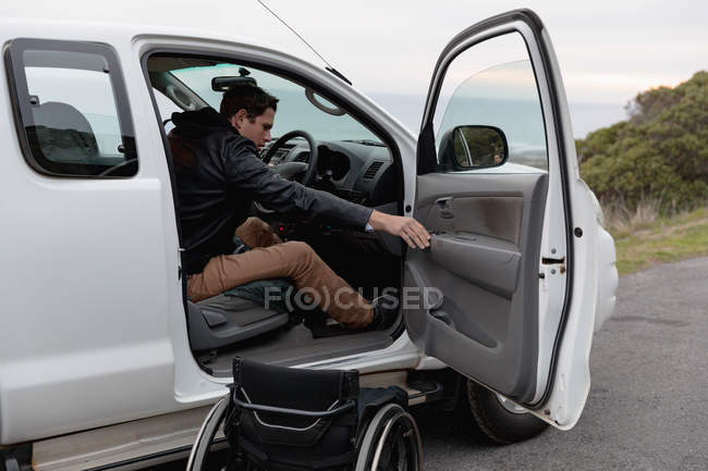 Vista lateral de un joven caucásico saliendo de una silla de ruedas y en su coche en un aparcamiento junto al mar - foto de stock